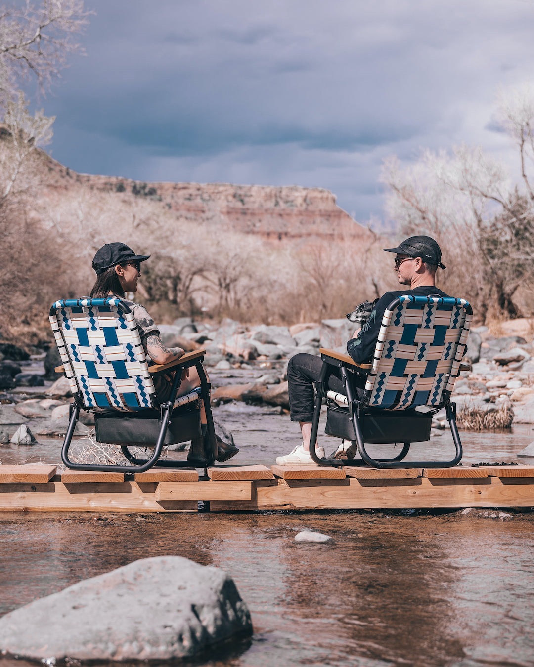 two people on camp chairs on a bridge over a small creek