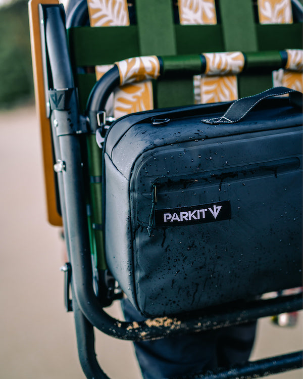 The back view of a voyager camping chair or beach chair when the folding chair is being carried by carrying straps on the users back like a backpack. You can see the cooler with it's storage pockets and sandy beach in the forground.