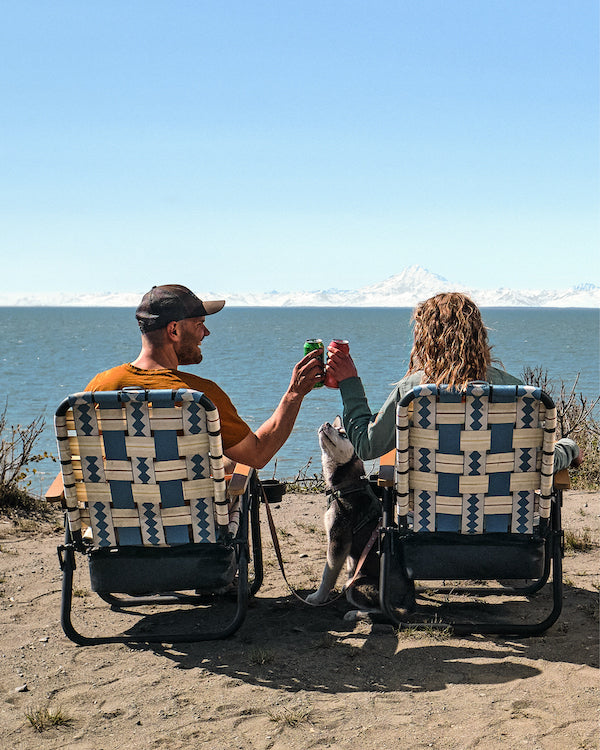 a couple sitting in the voyager camping chairs overlooking a beach from a cliff. they are cheersing two drinks that they had in the voyager chair cooler and their puppy husky sits between the two of tem as they take in the beatiful ocean view.