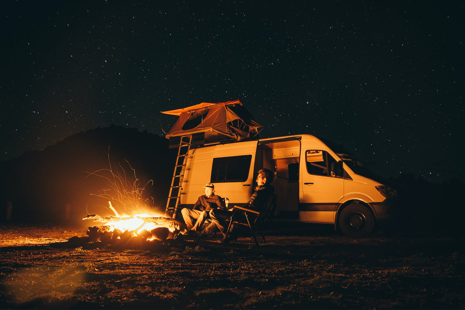 two friends sit in their voyager camping chairs while the moon and stars shine up above and the fire below keeps the two of them warm.